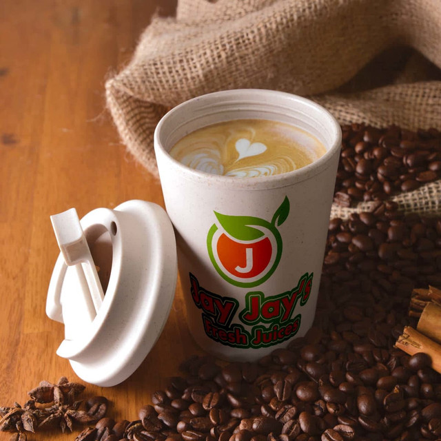 A reusable coffee cup in light beige with a comfort lid, featuring a logo, placed on a wooden surface with coffee beans.