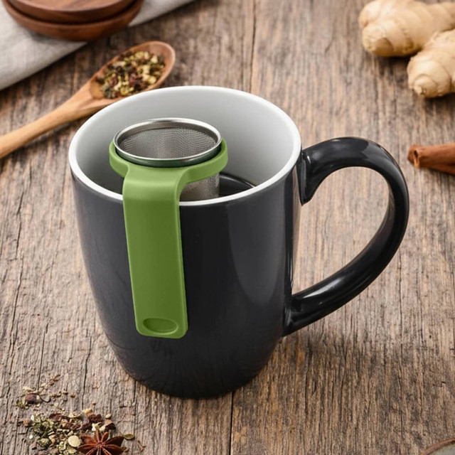 A drink bottle with a black exterior and green tea infuser sits in a mug, surrounded by loose tea and spices.
