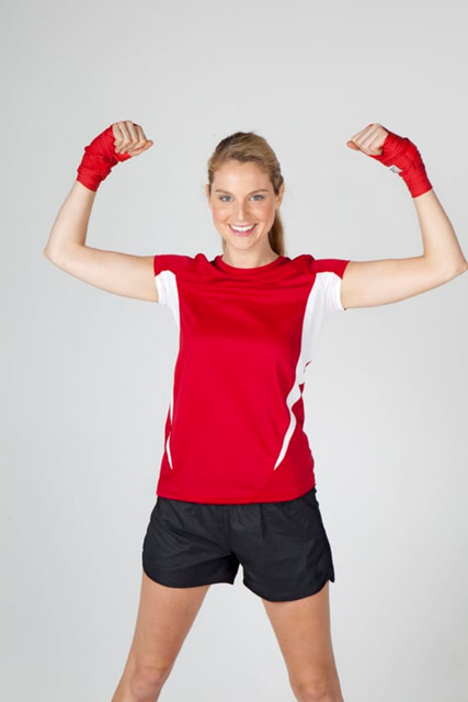 A woman in a red and white short-sleeved T-shirt flexes her arms, wearing black shorts and red hand wraps.