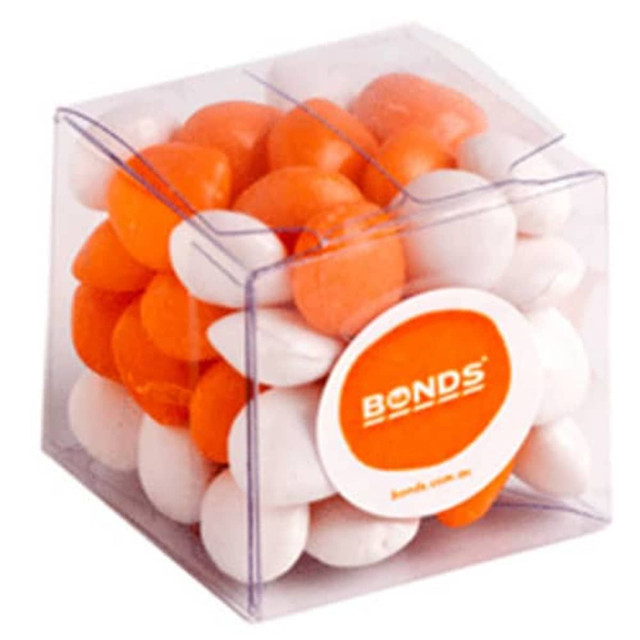 A cube-shaped container filled with orange and white chewy fruit candies. The container has a branding logo on it.
