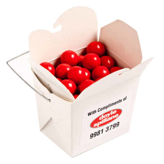 A white cardboard noodle box filled with bright orange chocolate drops. The box has a handle and a branding logo.