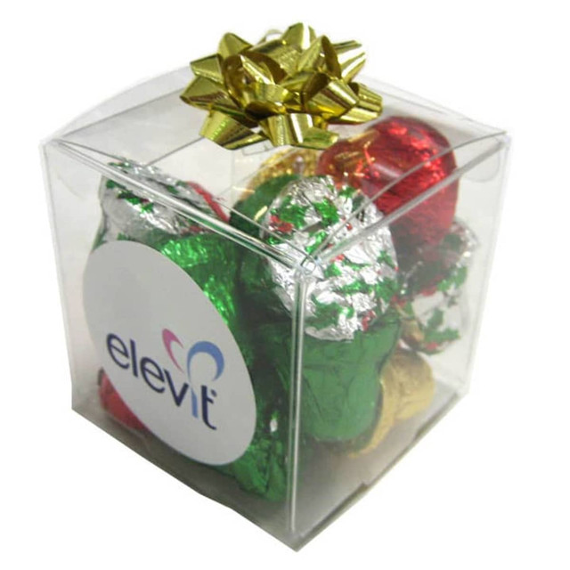 A clear cube filled with assorted Christmas chocolates wrapped in red, green, and silver foil, topped with a gold bow.