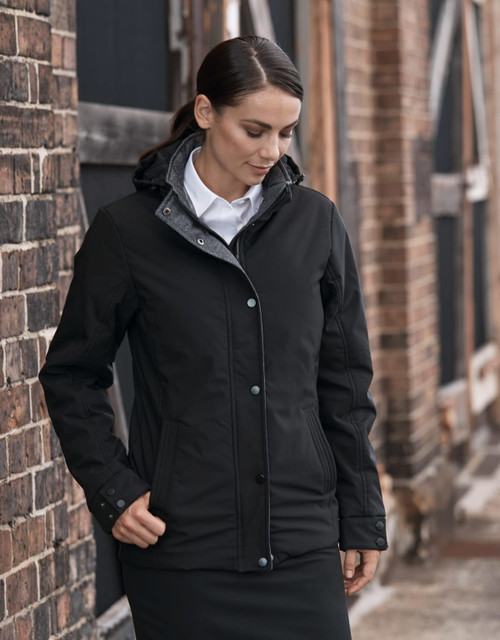 A black jacket with a hood, featuring a front zipper and buttons, worn by a model against a rustic brick backdrop.