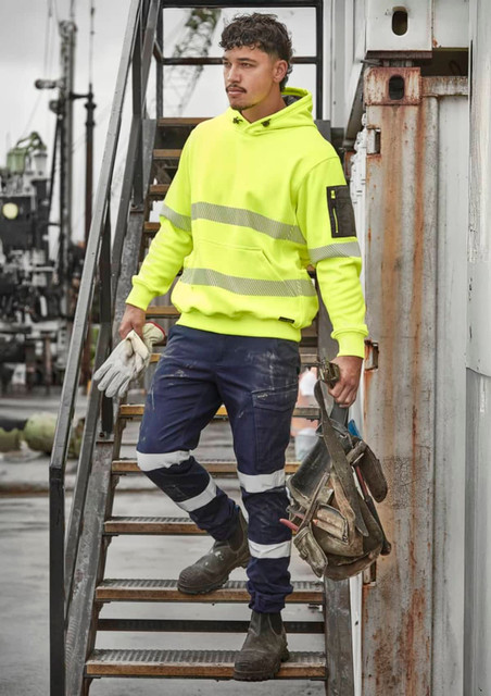 A man wearing a high-visibility yellow hoodie with reflective stripes and navy cargo pants, holding gloves and tools.