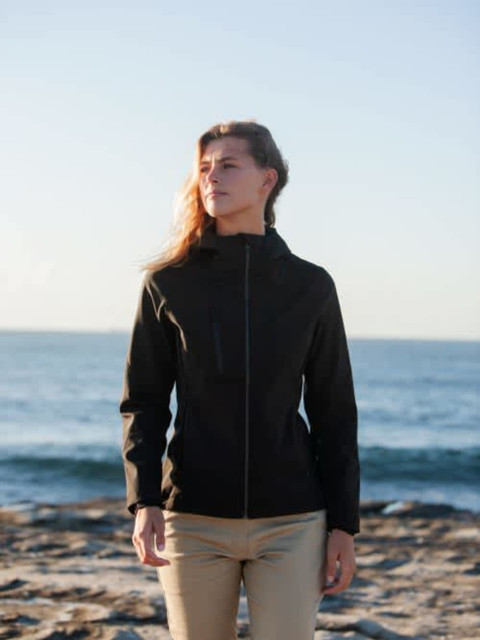 A woman stands by the beach in a black soft shell jacket, featuring a zip front and a sleek design.