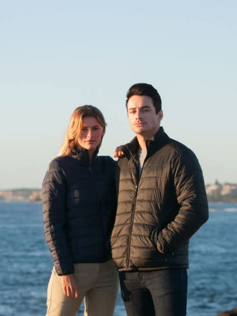A couple stands by the water wearing navy puffer jackets. The scene is coastal with a clear sky.