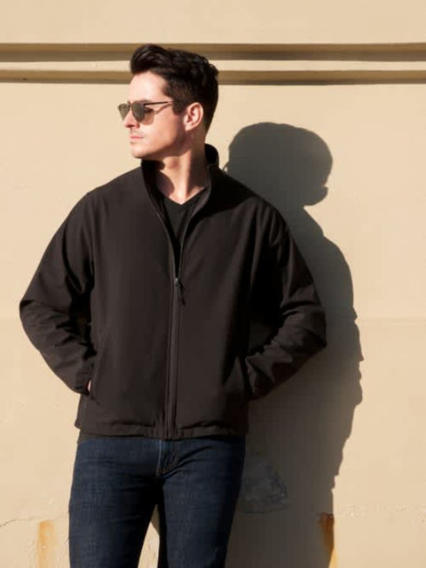 A man wearing a black soft shell jacket stands against a wall, with sunglasses and a shadow.