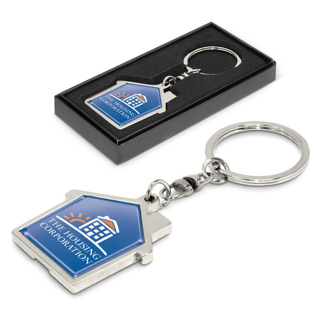 A house-shaped metal key ring in silver with a blue and white logo on a black presentation box.