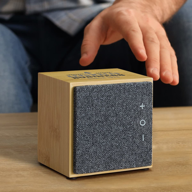 A square, bamboo Bluetooth speaker features a grey fabric front and volume controls, sitting on a wooden table.
