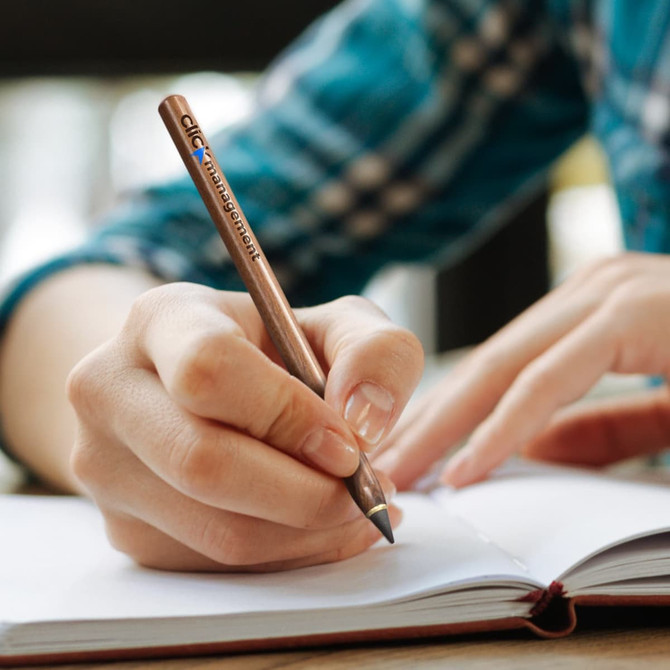 A metal pen with a bronze finish being held and used to write in an open notebook. The pen has a logo.