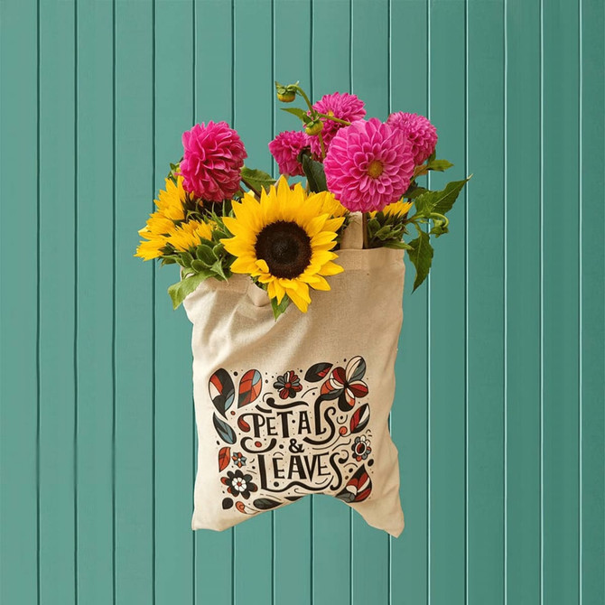 A cream canvas tote bag with a floral design, filled with vibrant sunflowers and pink dahlias.