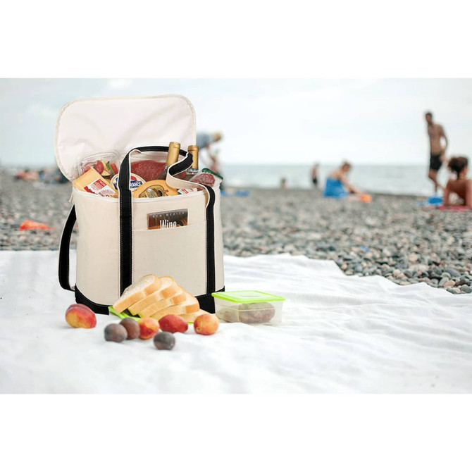 A cooler bag in cream with black straps, filled with drinks and food, set on a beach towel near people at the beach.