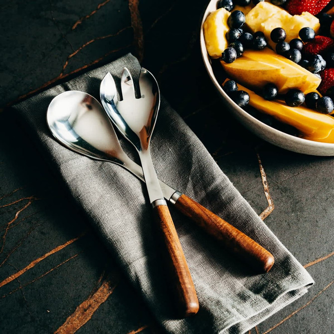 Stainless steel salad servers with wooden handles resting on a grey linen napkin, alongside a bowl of fresh fruit.