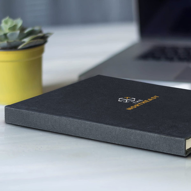 A recycled cotton hard cover notebook in black, placed on a desk next to a yellow plant pot and a laptop.