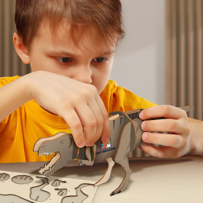 A child assembles a wooden Tyrannosaurus Rex model in shades of brown, surrounded by cardboard pieces on a table.