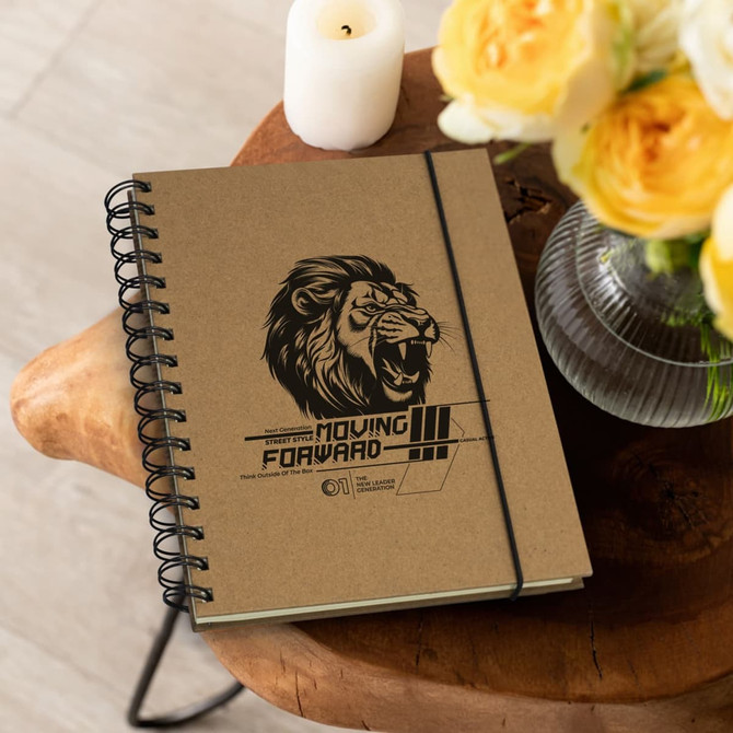 A brown spiral notebook with a lion graphic and a logo, placed on a wooden table beside yellow flowers and a candle.