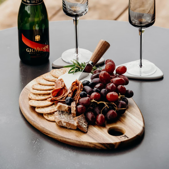 A wooden serving board with assorted crackers, grapes, cheese, and a knife, accompanied by champagne and glasses.