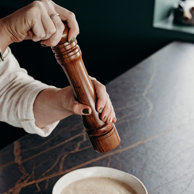 A wooden pepper mill being operated by a person, with a bowl of creamy mixture on a dark surface.