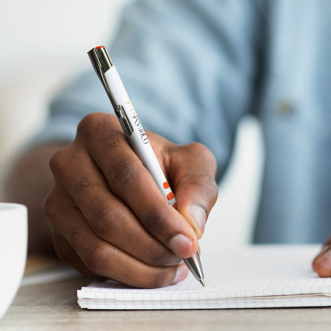 A hand holding a white barrel pen with metal accents, writing on a notepad. The pen features a logo.