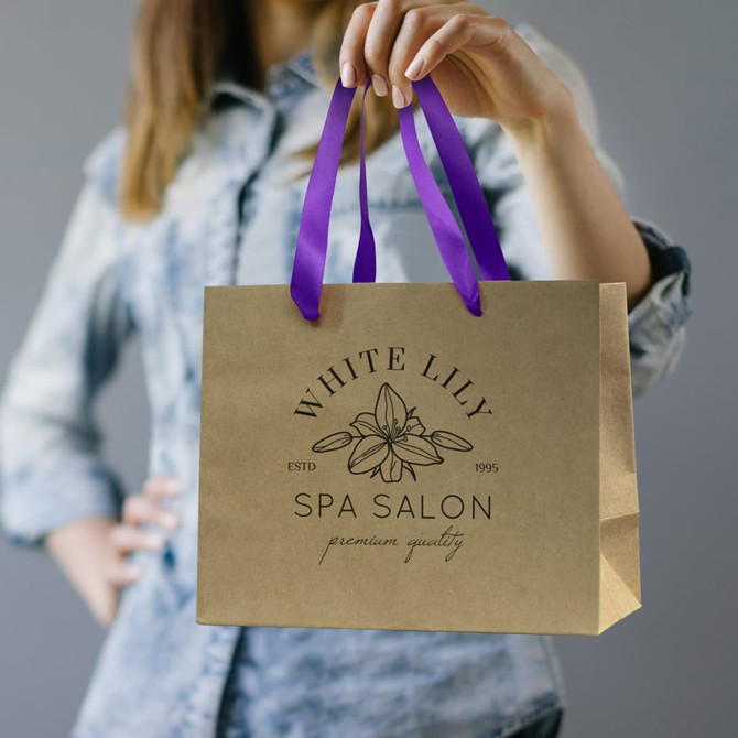 A medium brown paper bag with purple ribbon handles, featuring a logo on the front.