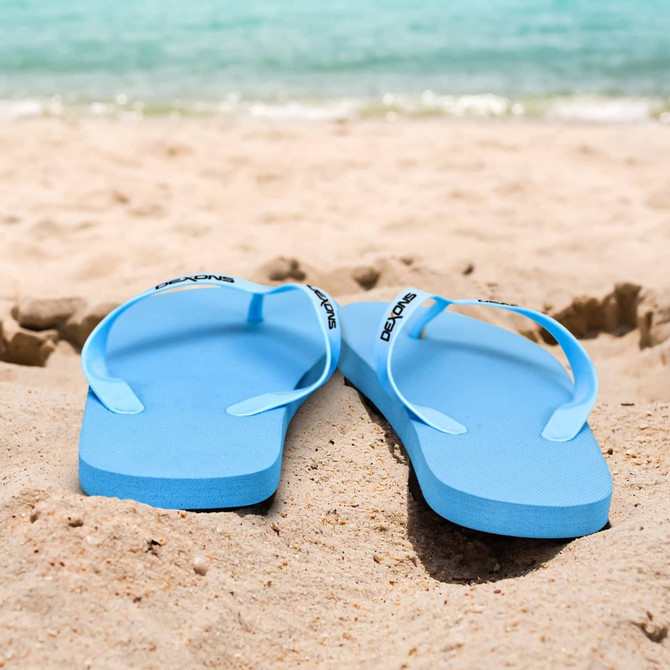 Blue flip flops resting on sandy beach with calm waters in the background. Features a logo on the strap.