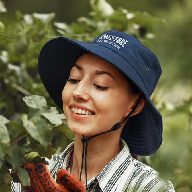 A navy Yuma Bucket Hat with a logo, worn by a smiling person among green foliage.