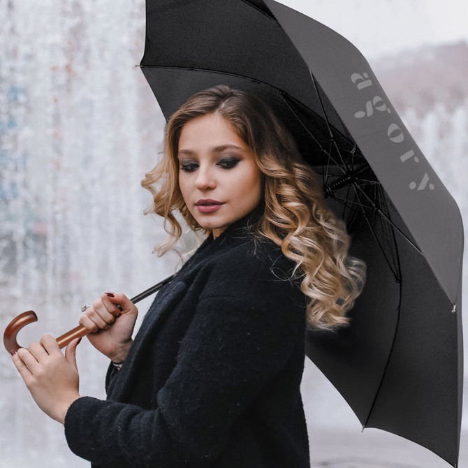 A black umbrella with a curved wooden handle, held by a person with long, wavy hair. The umbrella has a logo.