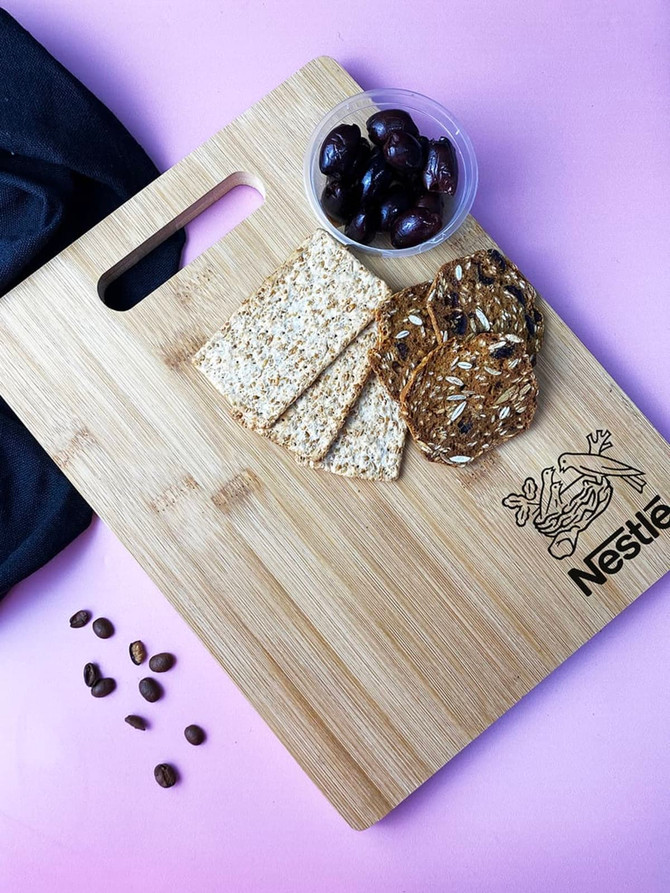 A bamboo chopping board featuring a logo, with crackers, seed biscuits, and a bowl of olives on a pink surface.