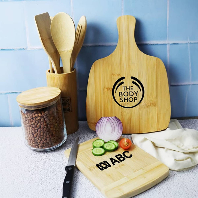 A bamboo cutting board in natural colour, alongside fresh vegetables, utensils, and a jar of ingredients. Features a logo.