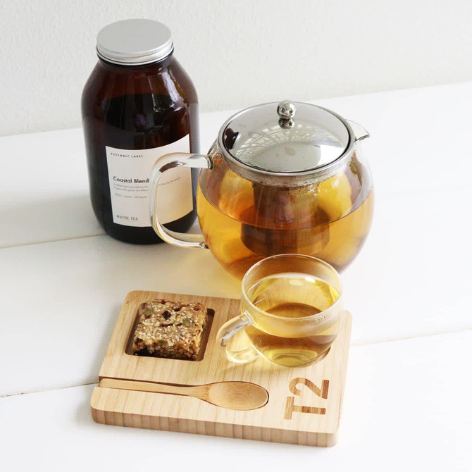 A glass teapot with a silver lid, a cup of tea, a wooden tray with a spoon, and a jar of tea leaves.