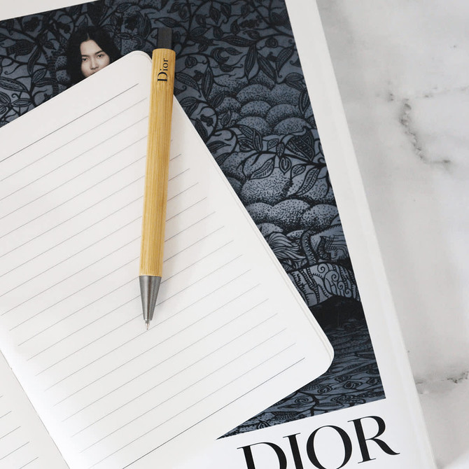 A bamboo pen with a metallic tip resting on a lined notebook, against a dark patterned background featuring a logo.