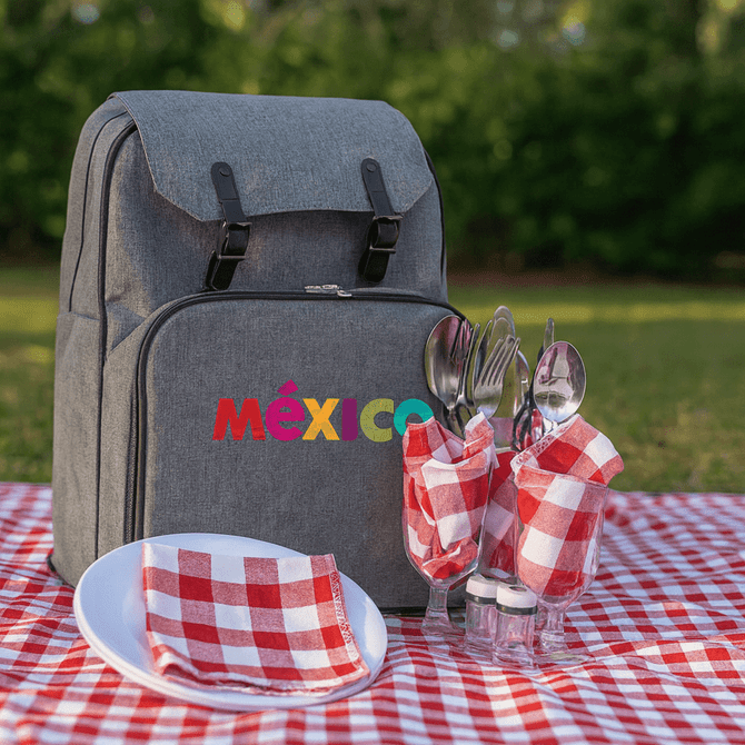 A grey picnic backpack rests on a red and white checkered picnic blanket, alongside utensils and napkins in glasses.