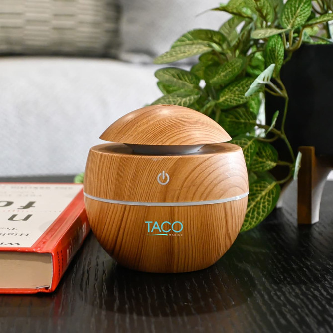 A wooden-patterned diffuser with a rounded design on a dark table, surrounded by greenery and a book.