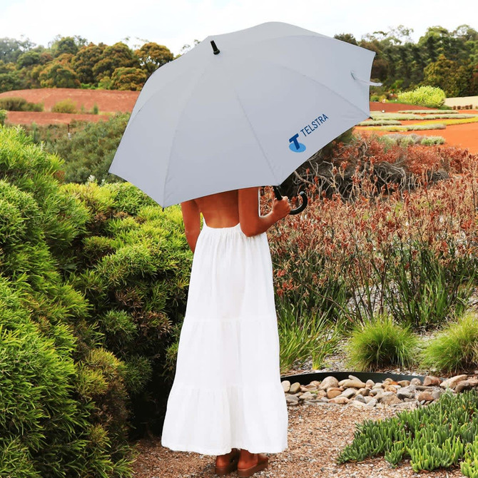 A grey umbrella with a logo is held by a person in a white dress, surrounded by garden scenery.