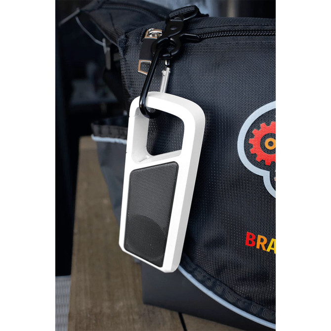 A white Bluetooth speaker with a mesh front and attachment clip, hanging from a bag. It features a logo on the bag.