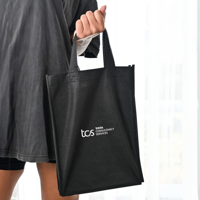 A black tote bag with two handles, featuring a logo on one side, held by a person.