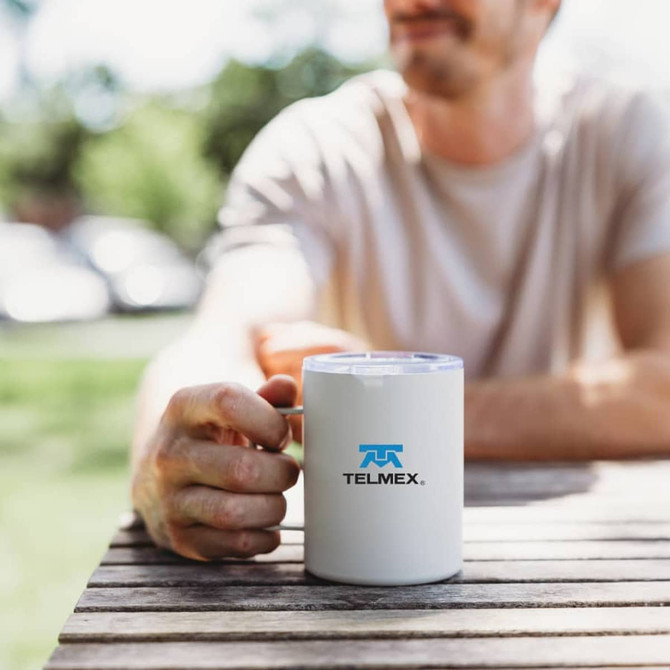 A travel mug featuring a white body and a blue logo, held by a person sitting at a table outdoors.