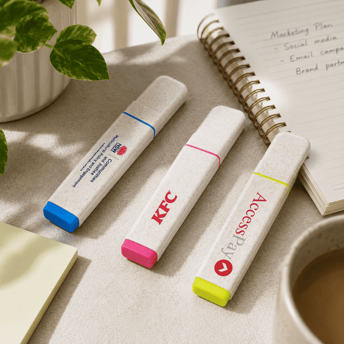 Three eco-friendly highlighters in blue, pink, and yellow on a desk next to a notepad and a plant.