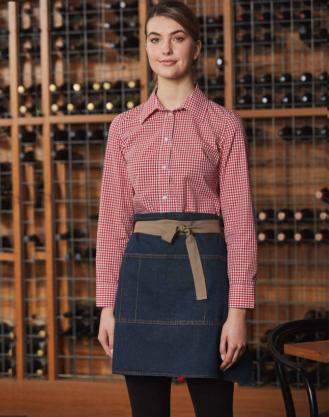 A model wears a denim half apron with a beige waistband, paired with a red checkered shirt against a wine rack backdrop.