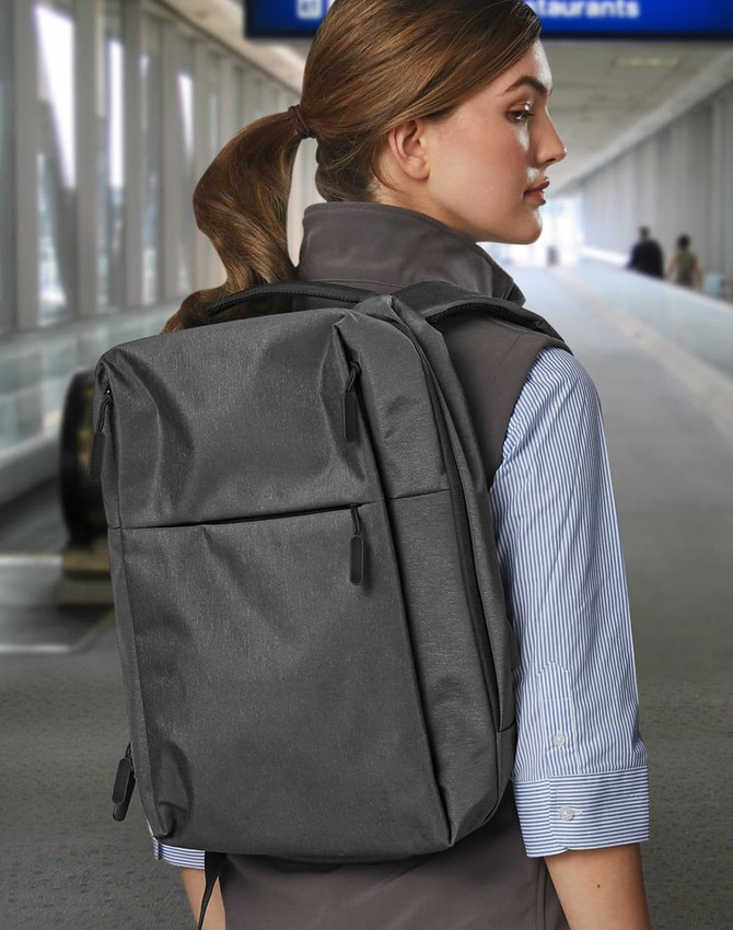 A grey backpack worn on the back of a person, set in a blurred airport environment. It features a logo.