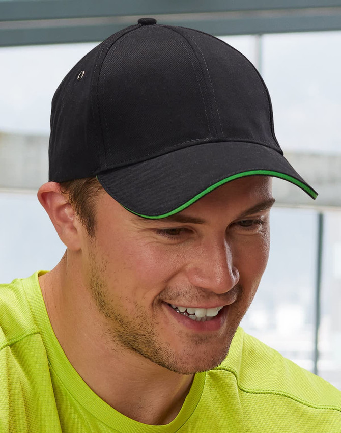 A black cap with a green peak, featuring a logo, worn by a smiling man in a bright yellow shirt.