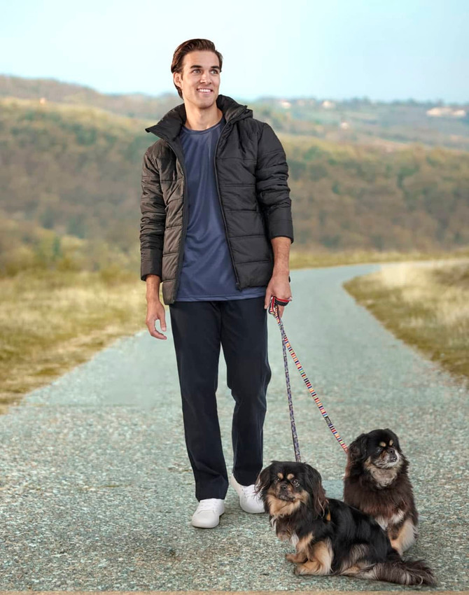 A man wearing a black sustainable parka jacket walks two dogs along a rural path, set against a hilly background.