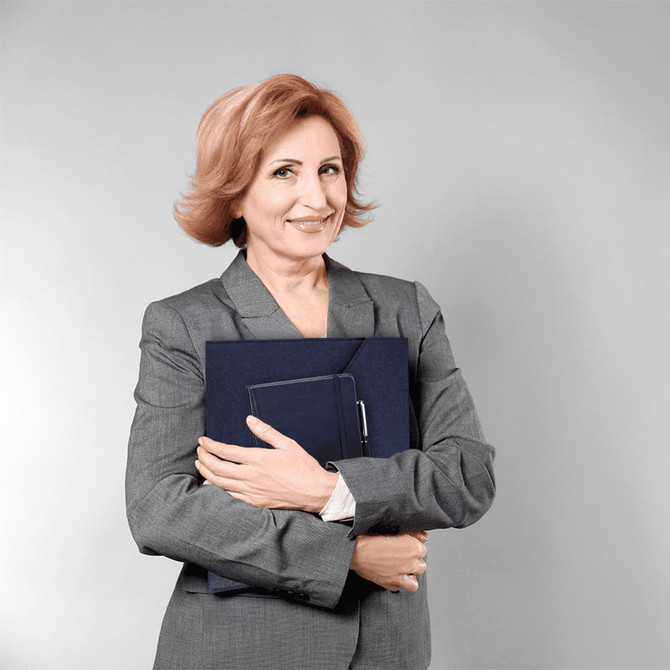 A woman in a grey suit smiles while holding a navy folder and a journal. The background is grey.