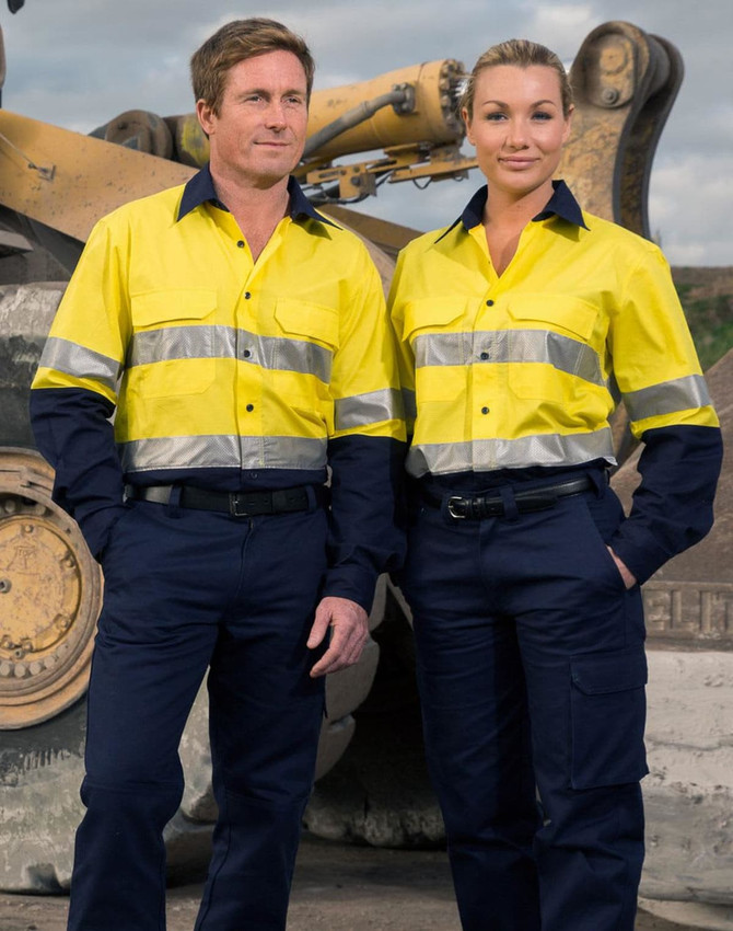 Two individuals wearing bright yellow long-sleeve shirts with reflective tape and navy pants, standing in an industrial setting.