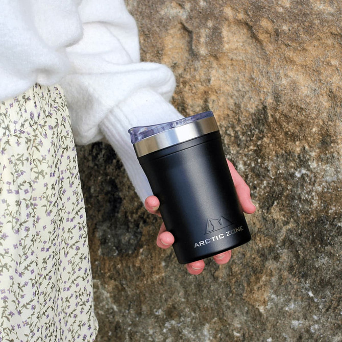 A drink bottle in matte black featuring a stainless steel lid, held by a person against a rocky background.