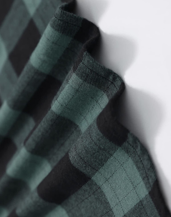 A close-up of a unisex flannel shirt in black and green plaid fabric, featuring a logo.