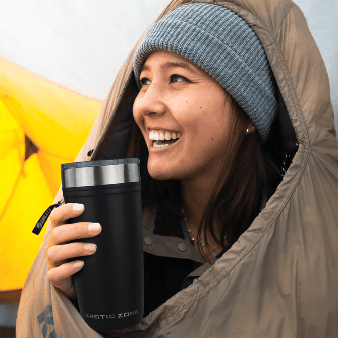 A drink bottle with a dark exterior and stainless steel top, held by a smiling woman in camping gear.