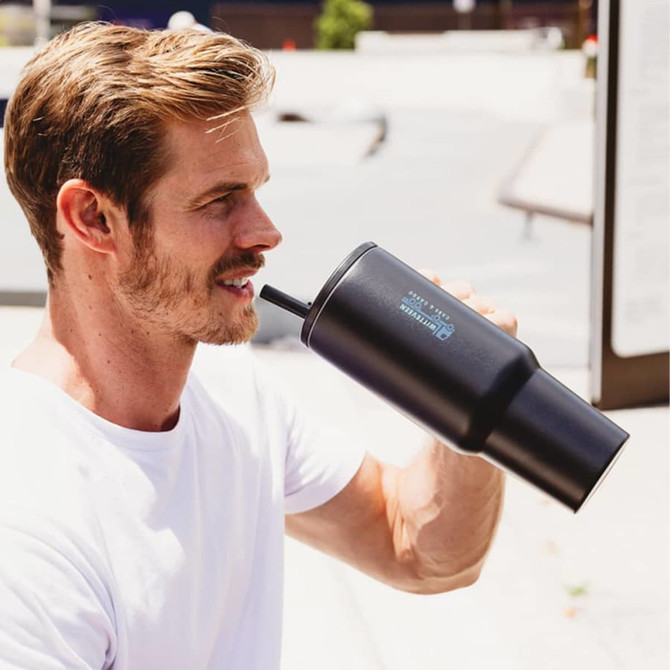 A drink bottle, the Rhino 1.2L Tumbler, in black with a sipping straw, held by a man outdoors.