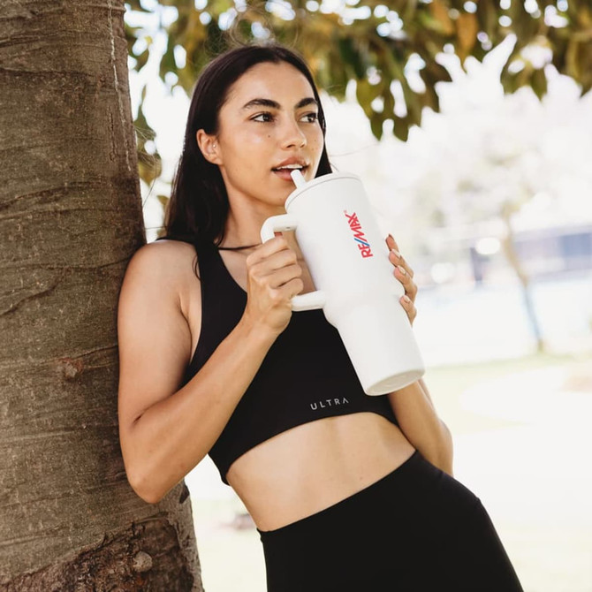 A drink bottle with a large white body and a straw, held by a person in a black sports outfit, leaning against a tree.