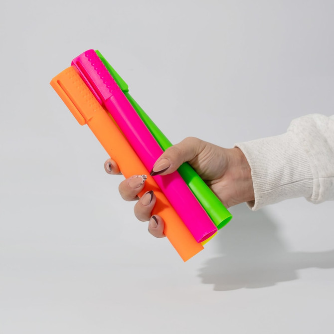 A hand holding three jumbo highlighters in vibrant neon pink, green, and orange colours, featuring a logo.